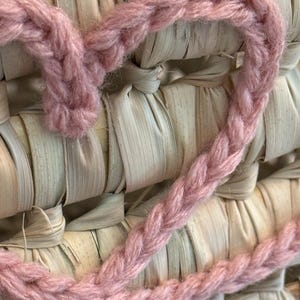 May include: A dusty pink crocheted heart shape made from thick yarn, layered over a woven background of light beige, flat, dried natural plant fibers arranged in a grid pattern with visible horizontal and vertical interlacing strips.