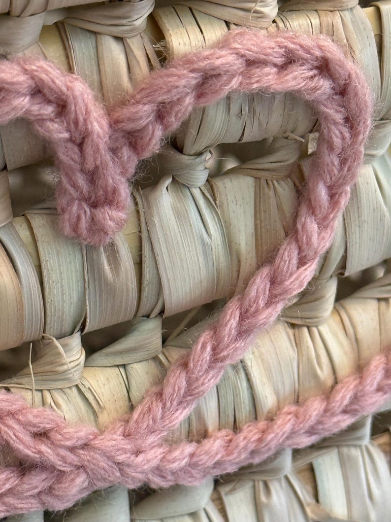 May include: A dusty pink crocheted heart shape made from thick yarn, layered over a woven background of light beige, flat, dried natural plant fibers arranged in a grid pattern with visible horizontal and vertical interlacing strips.