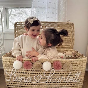 May include: A woven wicker basket with the names "Siena Scarlett" in white script. The basket is decorated with white, pink, and tan pom-poms. Two children are inside the basket, wearing matching beige sweaters.