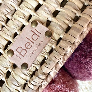 May include: A woven straw basket featuring a light tan leather patch embossed with the text Beldi Selection, secured by four brass-toned metal rivets. The basket is decorated with two large, soft pom-poms in shades of dusty rose and deep plum.