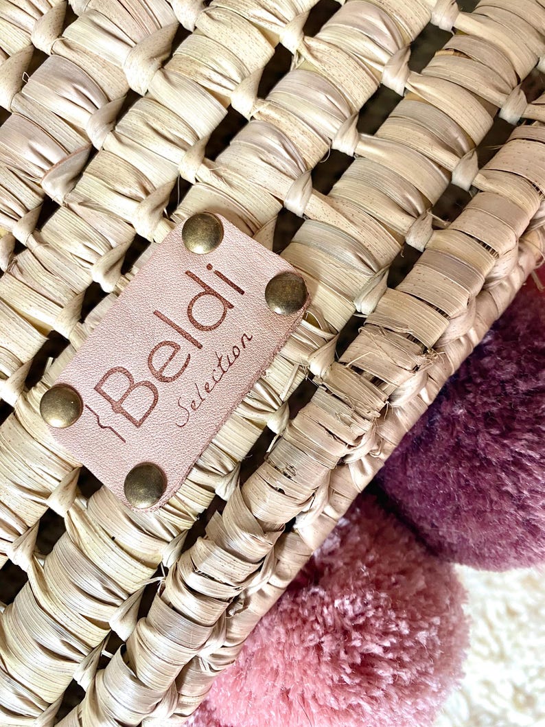 May include: A woven straw basket featuring a light tan leather patch embossed with the text Beldi Selection, secured by four brass-toned metal rivets. The basket is decorated with two large, soft pom-poms in shades of dusty rose and deep plum.