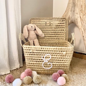 May include: A rectangular woven straw basket with a hinged lid and two side handles, featuring a white embroidered cursive letter E on the front. A plush beige rabbit toy sits inside the basket, and several small pom-poms in shades of pink, white, and tan are scattered around the base.