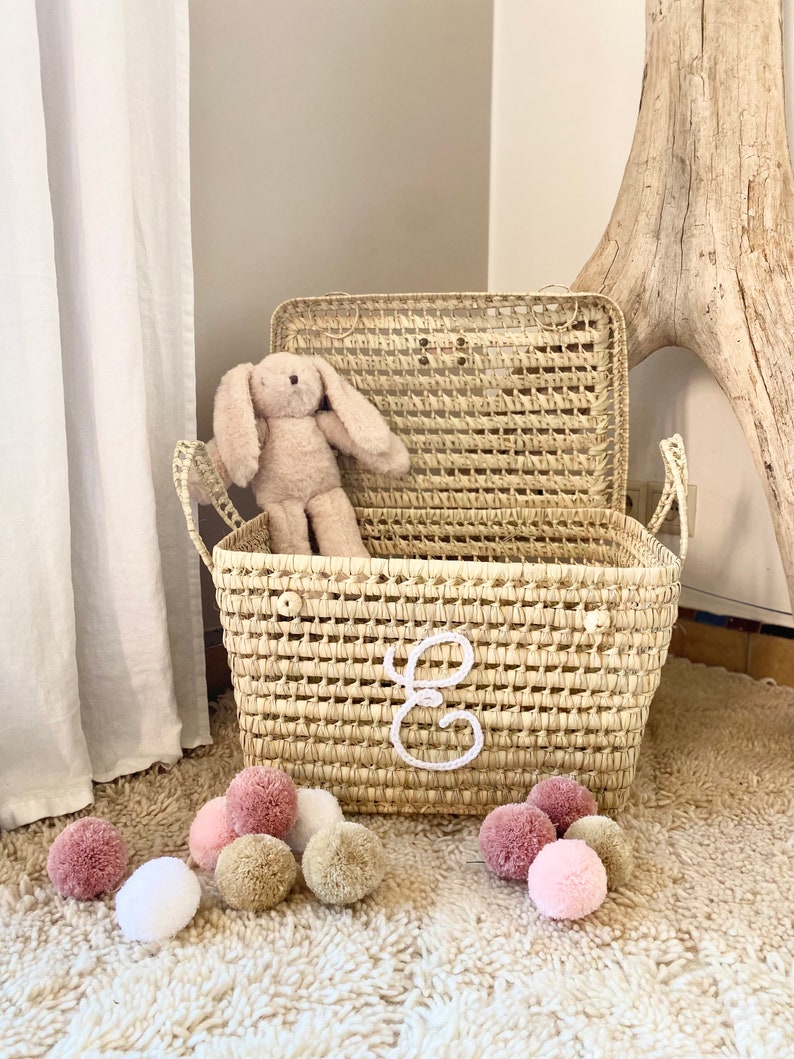 May include: A rectangular woven straw basket with a hinged lid and two side handles, featuring a white embroidered cursive letter E on the front. A plush beige rabbit toy sits inside the basket, and several small pom-poms in shades of pink, white, and tan are scattered around the base.