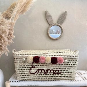 May include: A rectangular woven basket with a lid, featuring the name Emma embroidered in dark red script. The basket is decorated with a row of alternating beige, dark red, and dusty pink pom-poms along the front edge and two side handles.