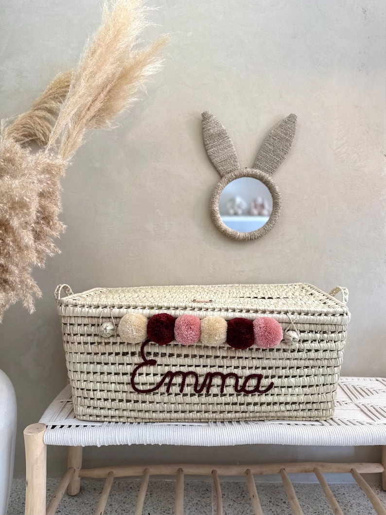May include: A rectangular woven basket with a lid, featuring the name Emma embroidered in dark red script. The basket is decorated with a row of alternating beige, dark red, and dusty pink pom-poms along the front edge and two side handles.