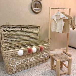 May include: A rectangular woven straw storage chest with a hinged lid, featuring the name Gemma Kate in white script, a row of five pom-poms in white, tan, and dusty rose, a wooden clothing rack with two garments, and a small wooden stool.