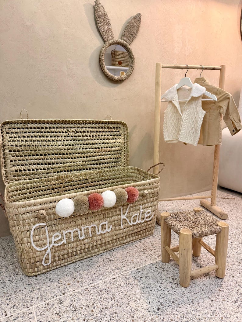 May include: A rectangular woven straw storage chest with a hinged lid, featuring the name Gemma Kate in white script, a row of five pom-poms in white, tan, and dusty rose, a wooden clothing rack with two garments, and a small wooden stool.