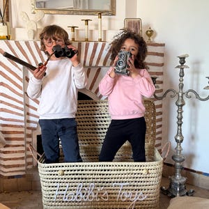 May include: A rectangular woven storage basket made of light-colored natural fibers featuring the white cursive text "Habibi's Toys" on the front. The basket has two side handles and a woven texture, providing a sturdy container for various items.