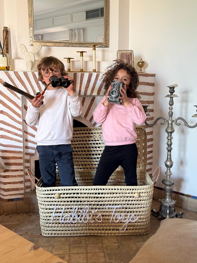 May include: A rectangular woven storage basket made of light-colored natural fibers featuring the white cursive text "Habibi's Toys" on the front. The basket has two side handles and a woven texture, providing a sturdy container for various items.