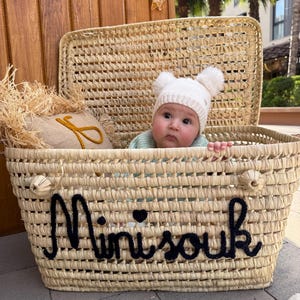 May include: A woven straw basket with a hinged lid featuring the black embroidered text Mini souk on the front. Inside the basket sits a baby wearing a white knit beanie with two pom-poms and a light green sweater next to a small burlap pillow.