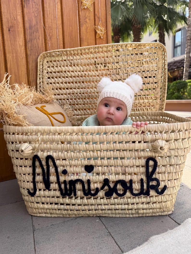May include: A woven straw basket with a hinged lid featuring the black embroidered text Mini souk on the front. Inside the basket sits a baby wearing a white knit beanie with two pom-poms and a light green sweater next to a small burlap pillow.