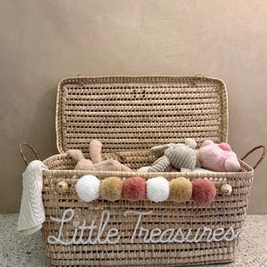 May include: A rectangular woven straw storage basket with a hinged lid and two side handles. The front features the white embroidered text Little Treasures and a row of six multi-colored pom-poms in white, tan, and dusty rose shades.