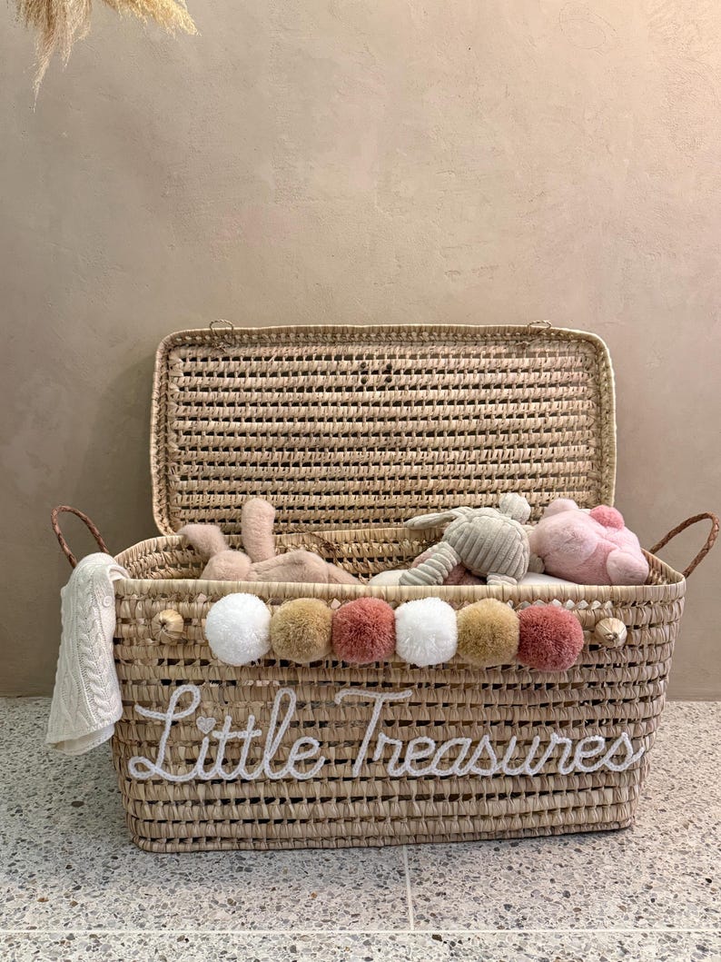 May include: A rectangular woven straw storage basket with a hinged lid and two side handles. The front features the white embroidered text Little Treasures and a row of six multi-colored pom-poms in white, tan, and dusty rose shades.