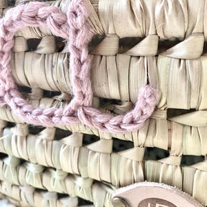 May include: A woven straw basket featuring a large, dusty pink crocheted letter J attached to the front. A small, oval-shaped light tan leather tag is secured with two brass-colored metal rivets and displays the letter B in a dark brown font.