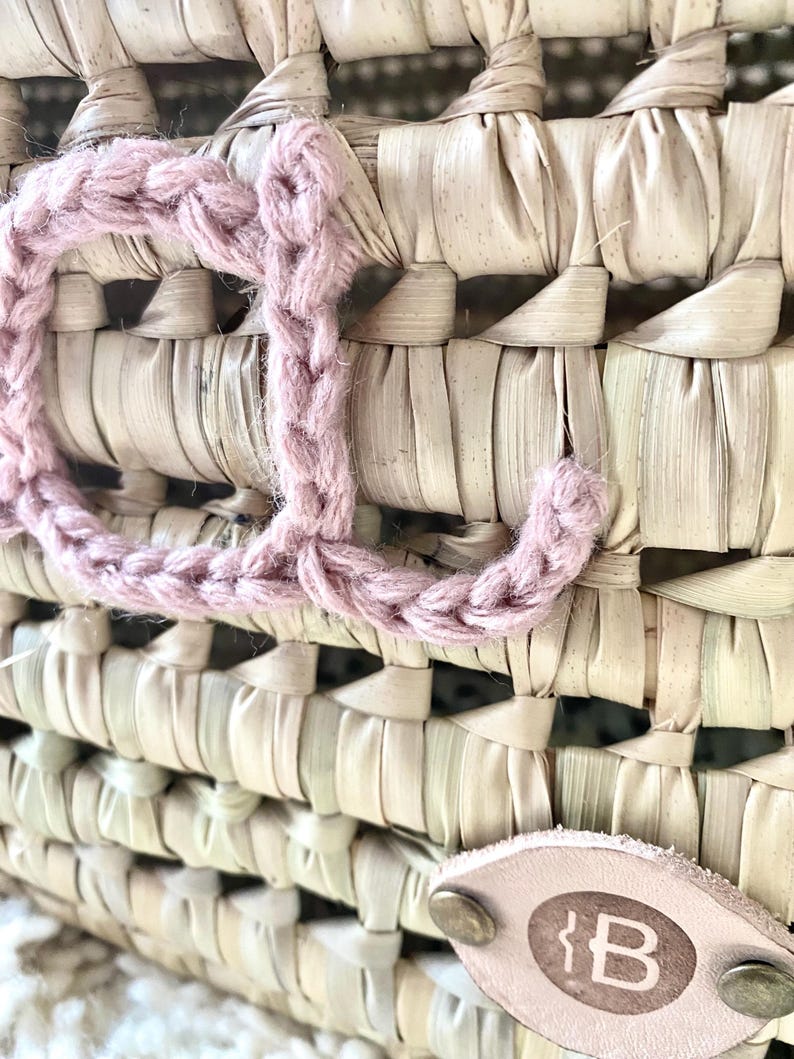 May include: A woven straw basket featuring a large, dusty pink crocheted letter J attached to the front. A small, oval-shaped light tan leather tag is secured with two brass-colored metal rivets and displays the letter B in a dark brown font.
