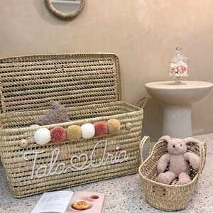 May include: A large rectangular woven straw storage basket with a lid, featuring a row of white, tan, and dusty pink pom-poms and the white cursive text "Tala & Aria". A smaller matching woven basket contains a plush rabbit toy.