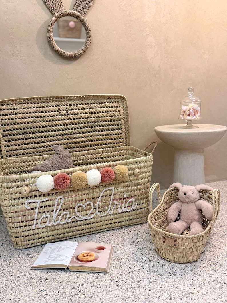May include: A large rectangular woven straw storage basket with a lid, featuring a row of white, tan, and dusty pink pom-poms and the white cursive text "Tala & Aria". A smaller matching woven basket contains a plush rabbit toy.