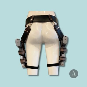 May include: A black leather gun belt and holster system with two silver and black pistols. The belt has brown leather accents and silver hardware. The holsters are attached to the belt and leg straps.