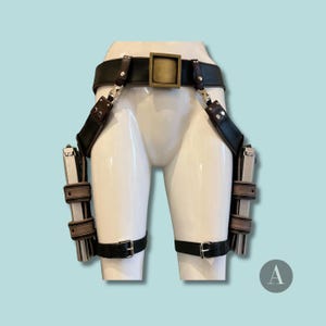 May include: Black leather gun holster with two silver and black pistols. The holster features a wide belt with a square buckle, suspenders, and leg straps. The background is a light blue.