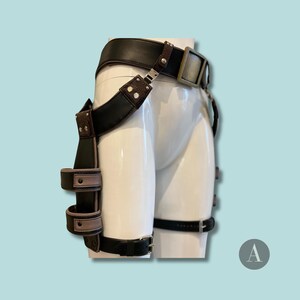 May include: A black and brown leather harness with a belt, leg straps, and attached accessories. The harness features a large rectangular buckle and is displayed on a white mannequin against a light blue background.
