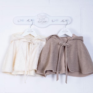 May include: Two hooded capes hanging on a white rack. The cape on the left is white with a lace pattern. The cape on the right is a light brown color. Both capes have a bow at the neck and are labeled "Little So & Sew".
