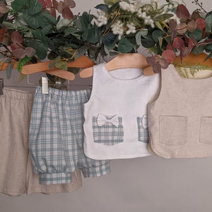 May include: Three pieces of children's clothing hanging on wooden hangers. A white sleeveless top with a plaid pocket and a white bow, a pair of blue and white plaid shorts, and a beige linen vest with two pockets.