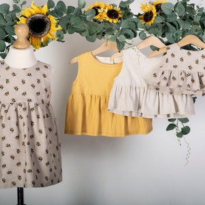 May include: Three sleeveless dresses for girls. One dress is beige with a floral print, one is yellow, and one is beige with a ruffle trim.