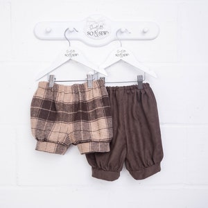 May include: Two pairs of baby bloomers hanging on white hangers. One pair is brown and tan plaid, the other is solid brown. The bloomers are made of soft fabric and have elastic waistbands and leg openings.