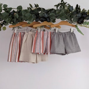 Op de afbeelding: Vier paar shorts hangen aan een waslijn. De shorts zijn in verschillende kleuren en patronen, waaronder rood-wit-blauwe strepen en effen grijs.