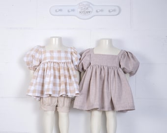 May include: Two toddler-sized dresses on mannequins. One dress is a beige and white gingham print with puffed sleeves. The other dress is a solid beige linen with puffed sleeves. Both dresses have a square neckline.