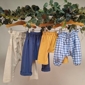 Può includere: Quattro paia di pantaloni per bambini appesi a uno stendino. I pantaloni sono realizzati in diversi tessuti e colori, tra cui beige, blu, giallo e gingham blu e bianco.
