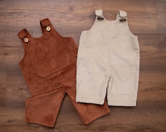 May include: Two pairs of corduroy overalls, one brown and one beige, laid out on a wooden surface.