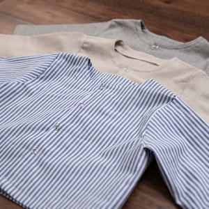 Op de afbeelding: Drie shirts met lange mouwen op elkaar gestapeld. Het bovenste shirt is grijs, het middelste shirt is beige en het onderste shirt is blauw-wit gestreept.