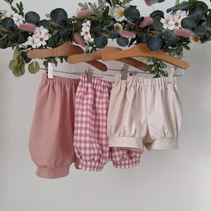 May include: Three pairs of baby bloomers hanging on wooden hangers. One pair is pink, one is white, and one is pink and white gingham.