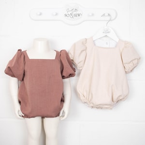 May include: Two baby onesies, one in a dusty rose color and one in a cream color, both with puffed sleeves and square necklines. The onesies are hanging on a white wooden hanger with the text "Sweet Little So & Sew" on it.