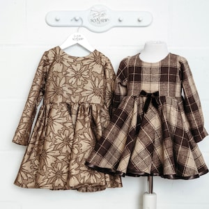 Könnte beinhalten: Zwei braune Kleider für Mädchen, eines mit Blumenmuster und das andere mit Karomuster. Beide Kleider haben lange Ärmel und hängen an Kleiderbügeln.