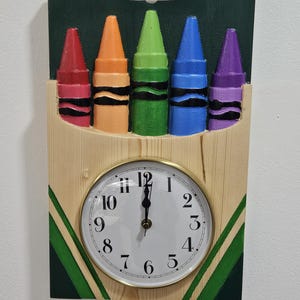 May include: A wooden clock featuring a design of five colorful crayons. The crayons are red, orange, green, blue, and purple, with black bands. The clock face is white with black numerals and hands, set in a gold-colored frame.