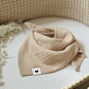 Könnte beinhalten: Beige Baby-Lätzchen aus Musselin mit einem kleinen weißen Etikett mit einem schwarzen Herz. Das Lätzchen ist gefaltet und liegt auf einer weißen Oberfläche mit kleinen grauen Punkten. Der Hintergrund umfasst einen geflochtenen Korb.