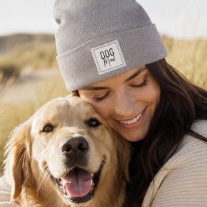 Könnte beinhalten: Eine hellgraue Strickmütze mit einem quadratischen weißen Aufnäher am vorderen Umschlag, auf dem der Text "DOG Mom" in schwarzen Buchstaben gedruckt ist. Die Mütze hat ein umgeschlagenes Design und eine strukturierte Strickoptik als Kopfbedeckung.