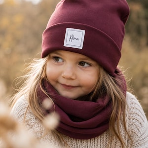 Könnte beinhalten: Eine tiefburgunderrote Beanie-Mütze mit umgeschlagenem Saum und einem weißen quadratischen Aufnäher, der den Schriftzug Alma in schwarzer Schreibschrift zeigt. Dazu gehört ein passender burgunderroter Schlauchschal aus glattem Stoff.