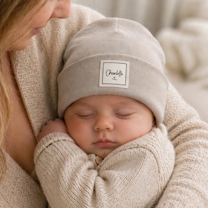 Könnte beinhalten: Ein schlafendes Baby trägt eine hellgraue Mütze mit dem Namen "Charlotte" auf einem weißen Etikett. Das Baby ist in einem beigefarbenen Pullover eingewickelt und wird von einer Person gehalten, die einen beigefarbenen Pullover trägt.