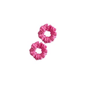May include: Two bright pink hair scrunchies with a smooth, satin-like finish. Each scrunchie features a gathered, circular fabric design with a central opening, showcasing a consistent vibrant pink color and a soft, reflective texture throughout.