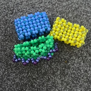 May include: Three beaded bracelets in blue, green, and yellow. The green bracelet has a purple trim.