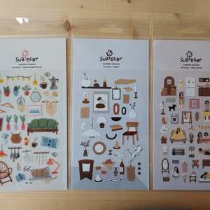 May include: Three sheets of stickers featuring illustrations of home decor, furniture, and other objects. The stickers are in a clear plastic bag and are labelled "Suatelier" with the text "Suatelier stickers no story, home sweet home".