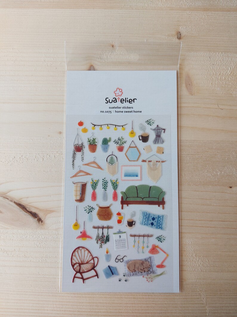 Pu&ograve; includere: Un foglio di adesivi trasparenti con illustrazioni di oggetti per la decorazione della casa, tra cui un divano, una lampada, un calendario, una pianta in vaso, un cesto, uno specchio, una caffettiera, un libro e un gatto. Gli adesivi sono etichettati "Sua elier, suatelier stickers, no.1075: home sweet home".