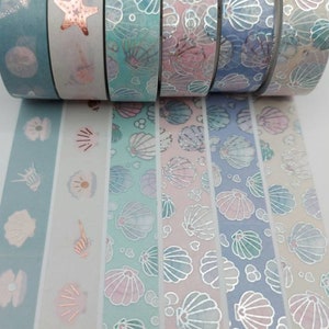 May include: A set of eight washi tapes featuring a seashell and starfish design. The tapes are in shades of blue, pink, and white with silver and gold accents.