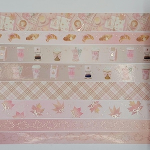 Puede incluir: Una colección de cintas washi decorativas con varios diseños. Las cintas presentan patrones de tazas de café, cruasanes, hojas de arce y cuadros en tonos rosa y dorado. Ideal para proyectos de scrapbooking y manualidades.