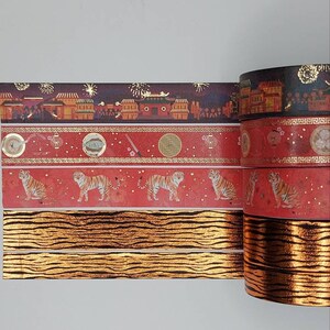May include: Four rolls of washi tape with gold accents. The first roll features a dark background with gold buildings and fireworks. The second roll has a red background with gold accents and a repeating pattern of tigers. The third and fourth rolls have a tiger stripe pattern with gold accents.