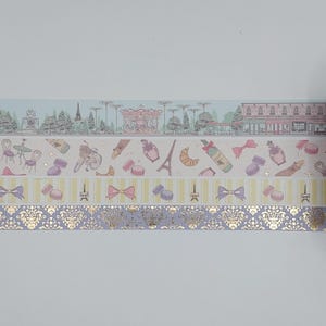 Puede incluir: Cinta adhesiva decorativa con temas parisinos. Los diseños incluyen la Torre Eiffel, macarons, lazos y un carrusel. La cinta tiene una variedad de patrones, incluyendo rayas y un diseño de damasco dorado. La paleta de colores incluye rosa pastel, morado y dorado.