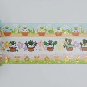 May include: Three rolls of washi tape with a floral and animal theme. The top roll has a light blue background with white animals in flower pots. The middle roll has a white background with pink flowers and green plants in flower pots. The bottom roll has a pink and green gradient background with gold flowers and bees.
